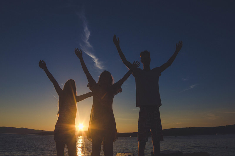 Three friends stood in the sunset by a lake with their hands in the air celebrating over 50s life cover